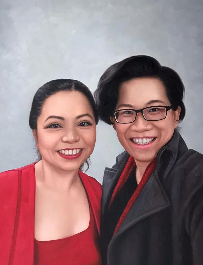 A man and woman smiling for a selfie against a neutral background. The woman has dark hair, is wearing a red top, and has makeup with red lipstick. The man has dark, styled hair, glasses, and is wearing a black jacket with a red lining.