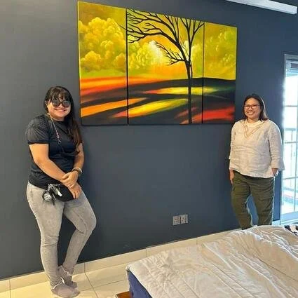 Two women standing in a room with a dark blue wall, a large colorful sunset landscape painting, and a bed with a white blanket.
