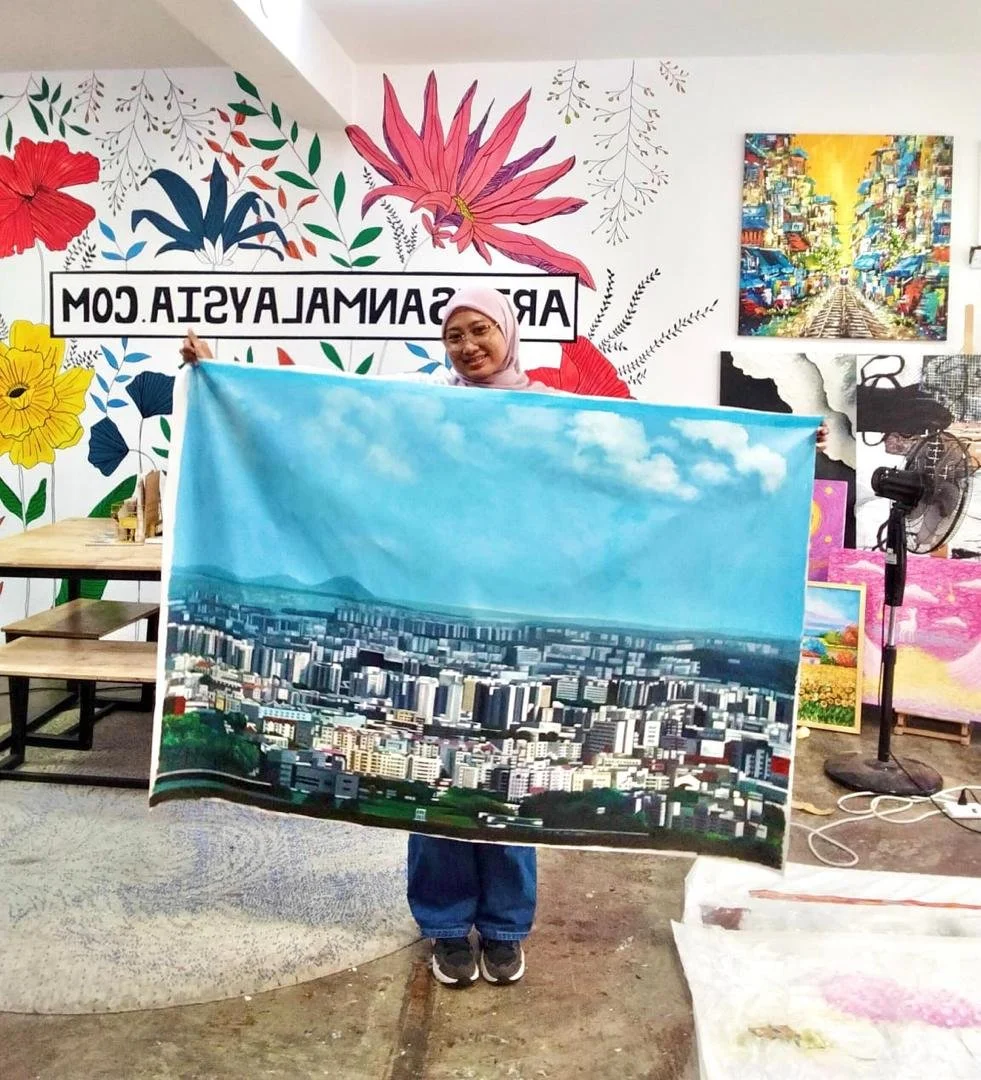 Woman in a pink headscarf holding a painting of a cityscape and sky in an art studio with colorful wall paintings.