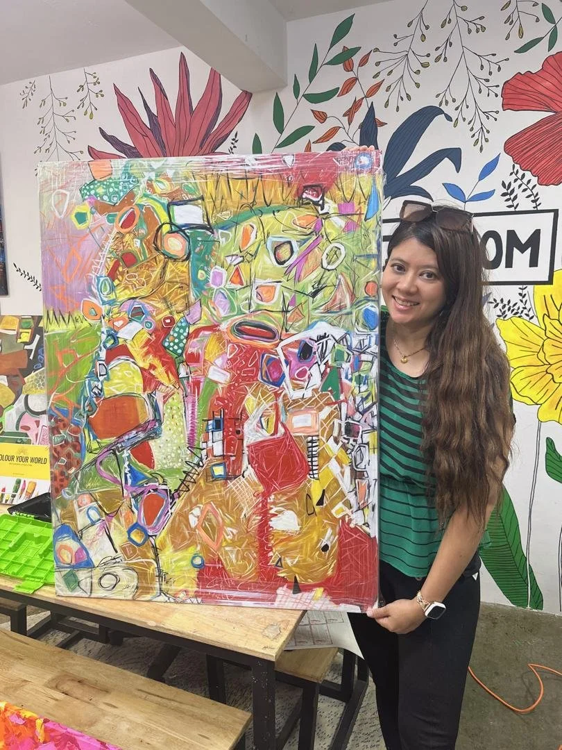 A woman with long wavy hair wearing a striped green and black shirt and glasses on her head, smiling and holding a colorful abstract painting in an art studio with floral illustrations on the wall.