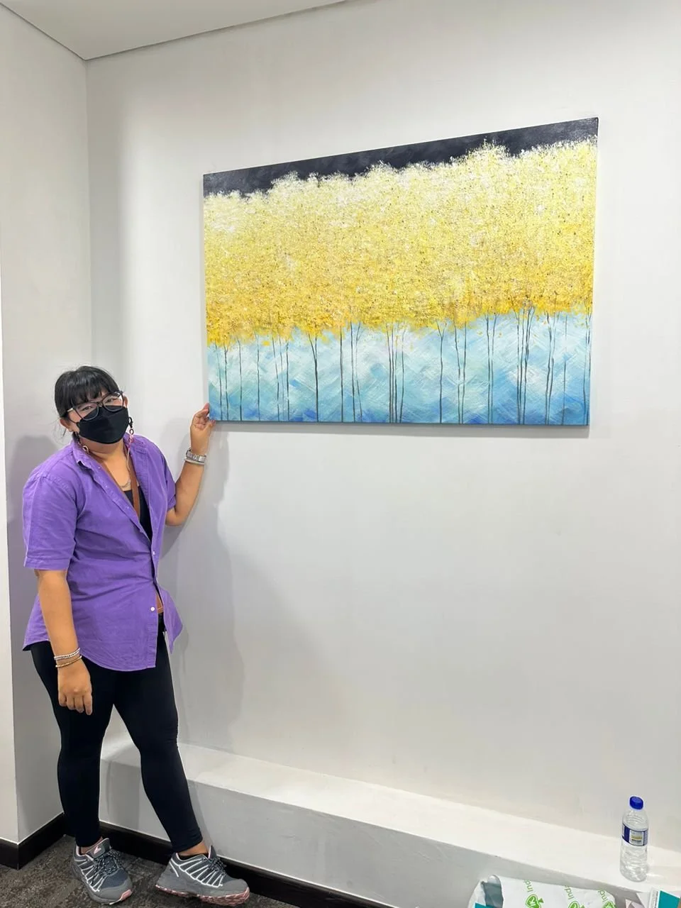 A woman wearing a purple shirt and a black face mask stands next to a large landscape painting of yellow trees with blue water underneath, hanging on a white wall.