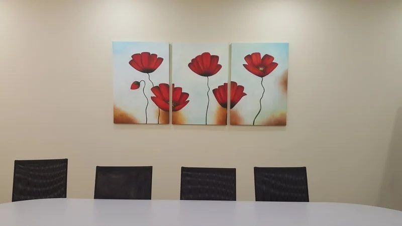 A set of four panels of a painting of red poppies on a white wall in a conference room with four black chairs and a white table.
