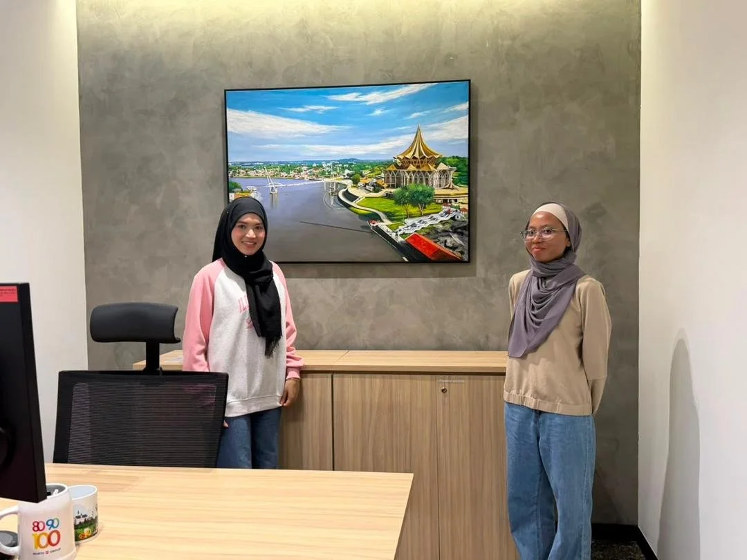 Two women wearing hijabs and casual clothes standing in an office beside a wooden cabinet, with a large screen displaying a scenic illustration of a cityscape and a temple in the background.