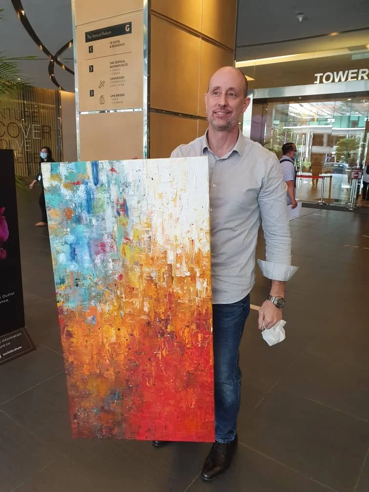 A man in a light gray button-up shirt and blue jeans holding an abstract painting with vibrant orange, red, yellow, blue, and white colors in a modern indoor space with glass and metal architecture.