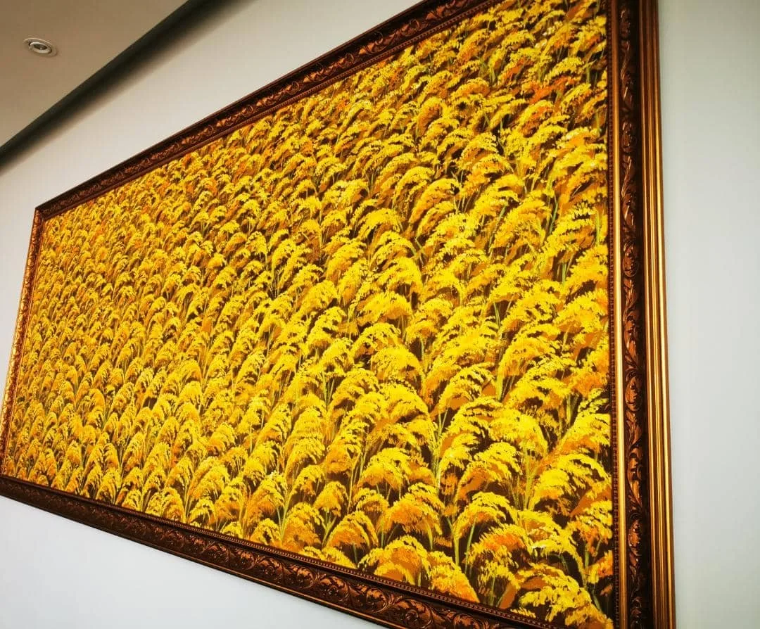 Painting of a yellow floral field in a decorative brown frame hanging on a white wall.