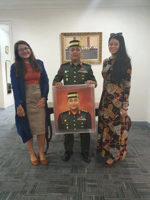 A man in a military uniform holding a framed portrait of himself, flanked by two women in stylish dresses, standing in a room with framed art on the wall.