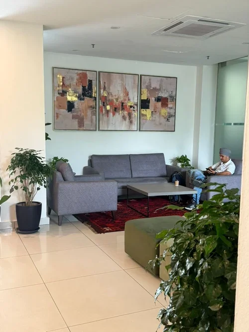 A modern waiting area with gray sofas, green plants, a red patterned rug, and three abstract paintings on the wall. A man is seated on the right side, reading or using a device.