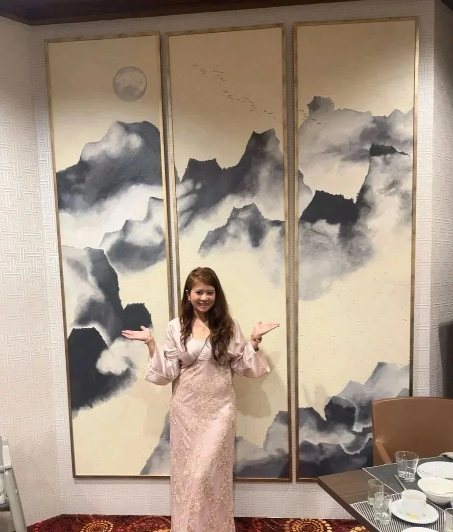A woman in a pink traditional dress standing in front of a triptych painting of mountains and a moon on a wall in a restaurant or dining area.
