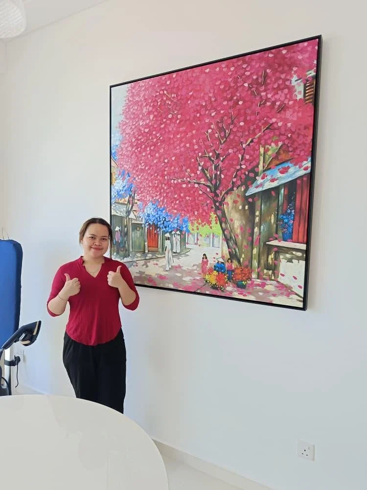 A woman standing next to a large colorful painting of a pink and blue flowering tree and traditional buildings, giving a thumbs-up.