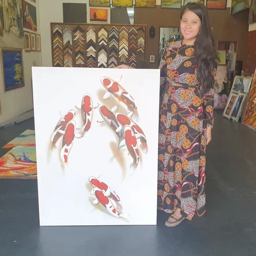 A woman with long black hair, wearing a colorful patterned dress, stands next to a large white artwork depicting koi fish. She is smiling and standing in an art gallery surrounded by various paintings and framed artwork.