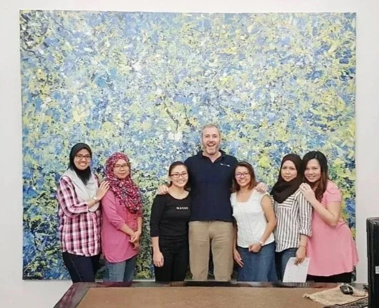 Group of seven people, including a man and six women, standing in front of a large colorful abstract painting, smiling for the photo.