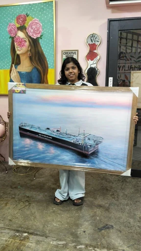 A girl holding a framed painting of a ship floats on water, standing indoors with colorful artwork and mirrors on the walls behind her.