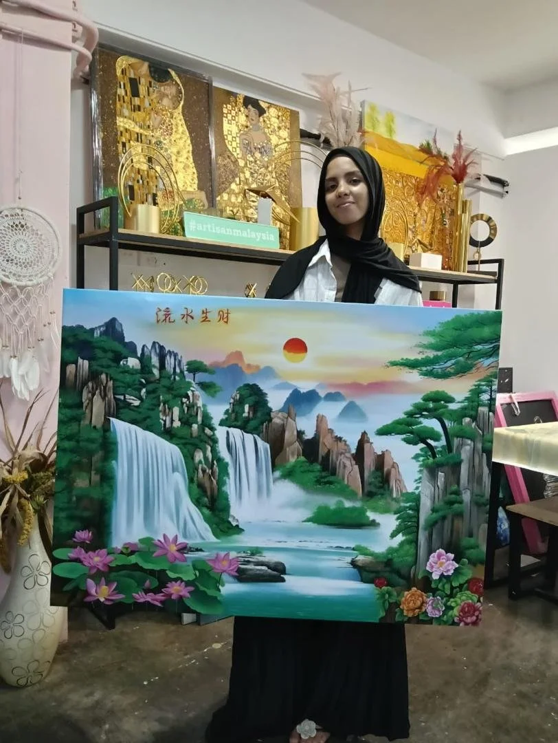 Young woman in hijab holding a landscape painting of waterfalls, mountains, trees, and a sunset, with Chinese characters in the top left corner, in an indoor art studio with decorative art pieces and a dreamcatcher on the wall.