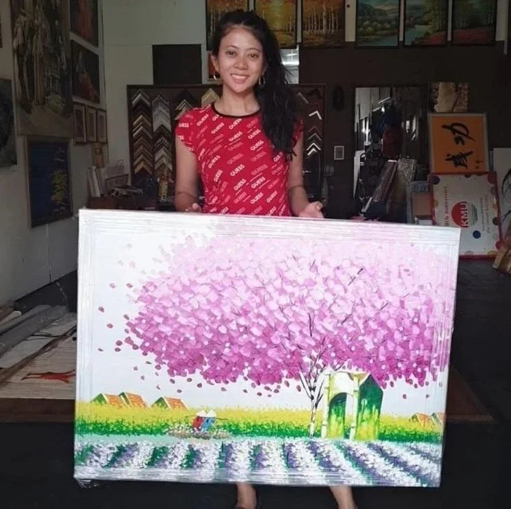 Woman smiling and holding a colorful painting of a pink blossoming tree, green fields, and a small house in an art studio.