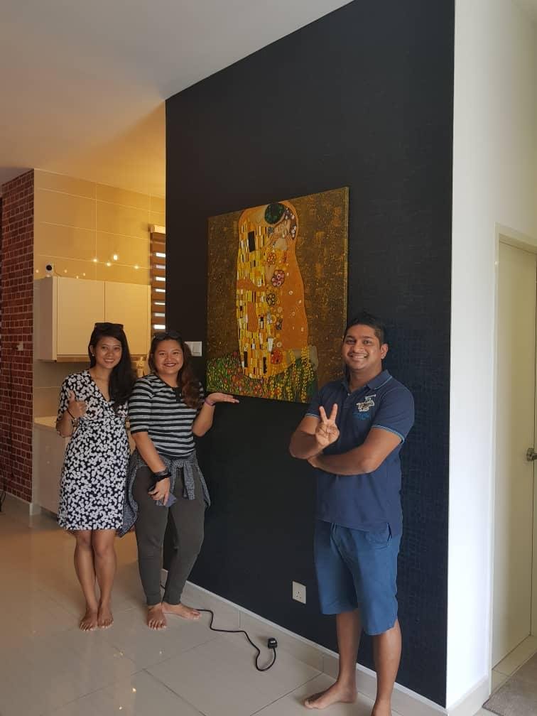 Three people standing in a room near a painting of a woman and a child, with the woman and child embracing. They are smiling, and one person holds up a peace sign. The room has a dark accent wall and a kitchen area in the background.