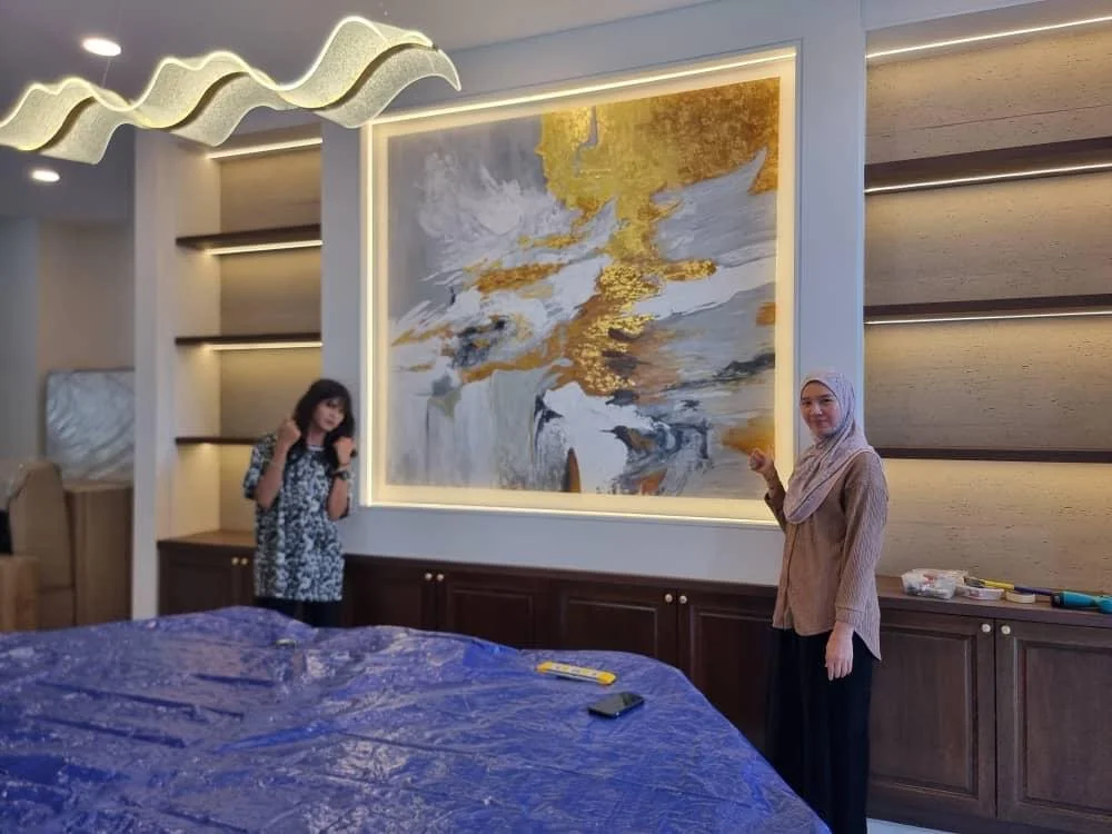 Two women in a modern room standing in front of a large abstract painting with white, gold, gray, and black colors. One woman is dressed in a floral dress and the other in a hijab and beige top. There are shelves and a large piece of wrapped furnitur