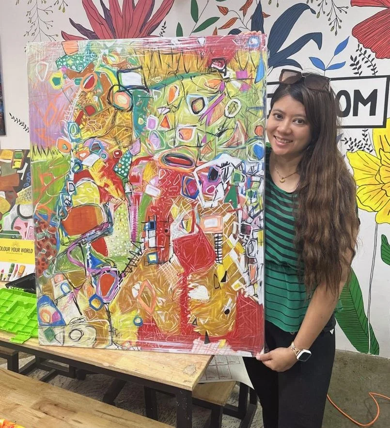 A young woman with long wavy hair smiling and holding an abstract colorful painting on a canvas, with a background of floral wall art.