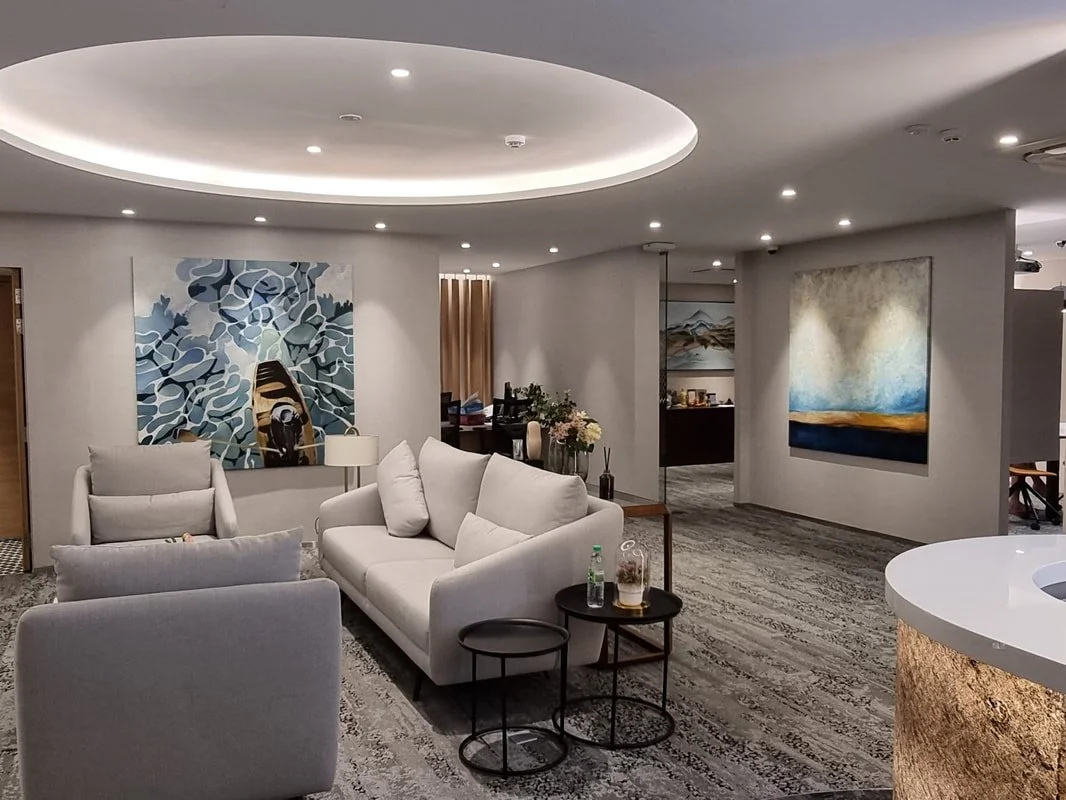 Modern hotel lobby with white sofas, armchairs, abstract paintings on the walls, and a circular ceiling light feature.