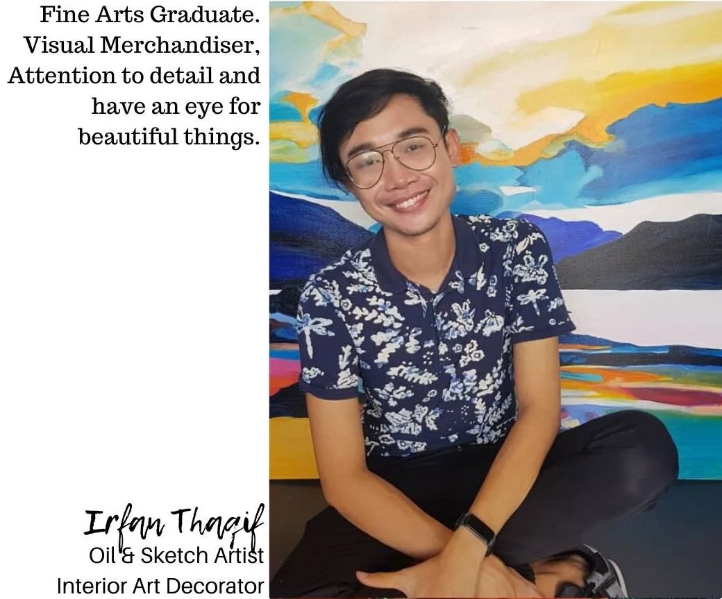 A young man with short black hair, glasses, and a smile, sitting cross-legged in front of a colorful landscape painting. He is wearing a blue shirt with white floral patterns and black pants.