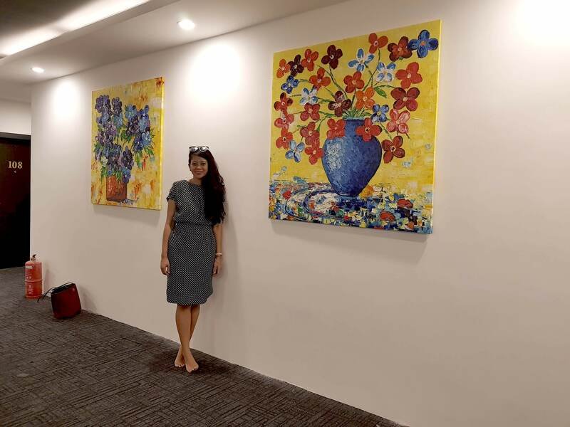 A woman in a polka dot dress standing next to two large colorful floral paintings on a white wall in an art gallery or hallway.