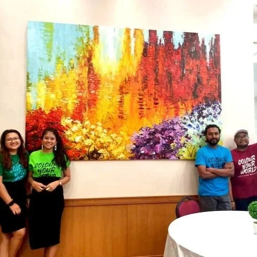 Four people standing in front of a large colorful abstract painting in a room with wood-paneled walls and a round table with a small plant in a white vase.