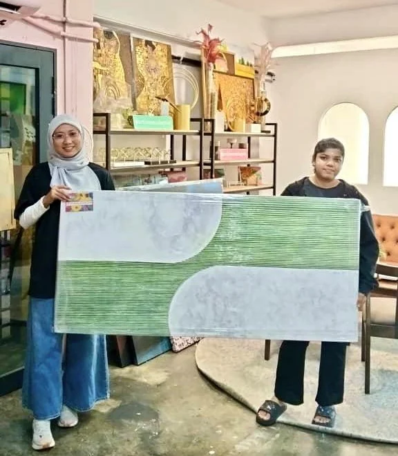 Two women standing indoors holding a large rectangular board with a green and white pattern. The woman on the left is wearing a white headscarf, black top, and blue pants. The woman on the right is dressed in black. They are in a room decorated with 