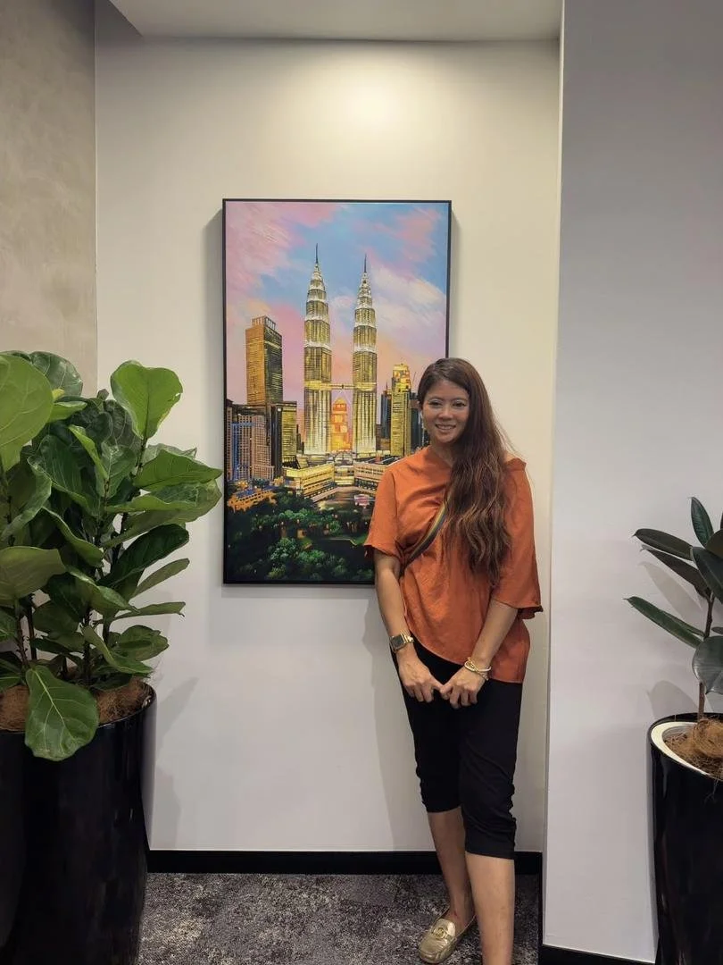 A woman with long brown hair wearing an orange top, black pants, and gold shoes standing indoors near a painting of the Petronas Towers in Kuala Lumpur, Malaysia. There are large potted plants on each side.