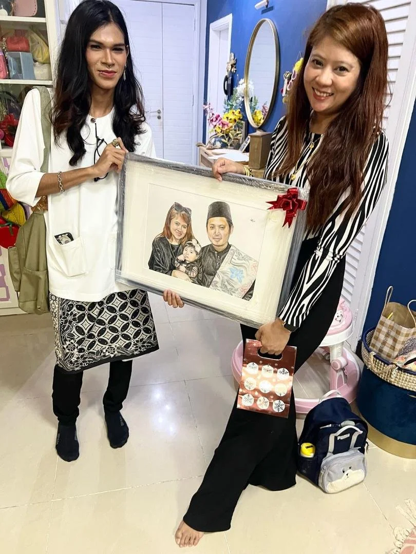 Two women are smiling and holding a framed drawing of a family portrait. One woman has long black hair, and the other has long reddish-brown hair. The woman with reddish hair is also holding a small box of cupcakes and is barefoot. The scene appears 