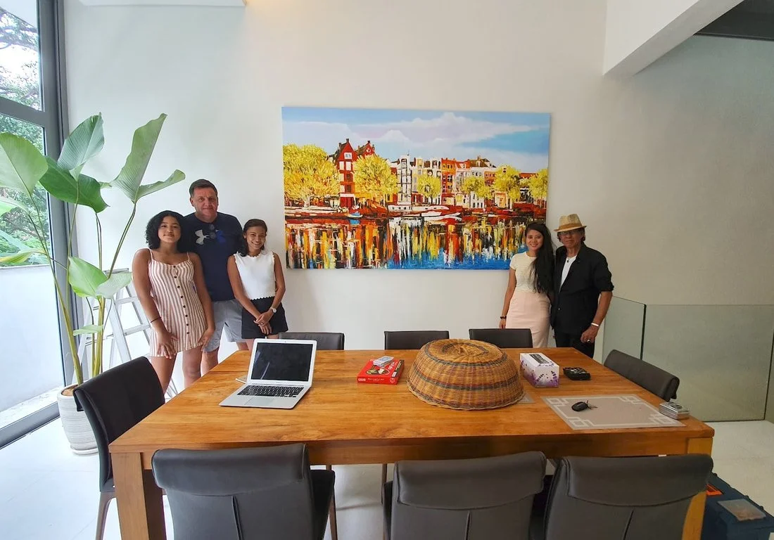Five people standing in a modern conference room with a large colorful painting of a European cityscape on the white wall behind them. A laptop, tissue box, and other items are on the wooden table in the foreground.