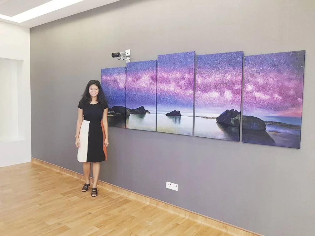 A woman standing next to a five-panel landscape painting of a starry night sky over water and rocks in a modern room with wooden floors.