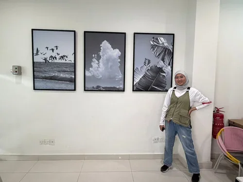 A person wearing a white headscarf, glasses, white shirt, green vest, and jeans standing in front of three framed black-and-white nature-themed photographs on a white wall, with a pink chair and a fire extinguisher nearby.