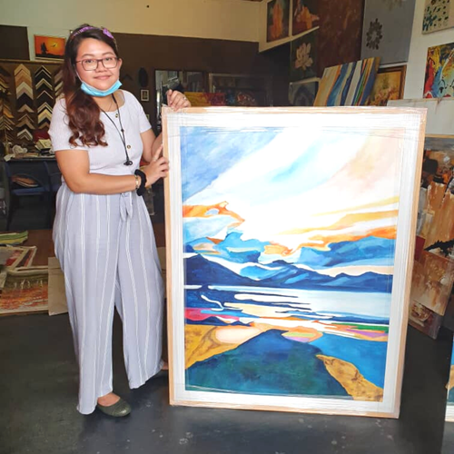 A young woman wearing glasses, a face mask hanging below her chin, and a headband, standing next to a colorful painting of a seascape in an art gallery or studio, surrounded by other artworks and frames.