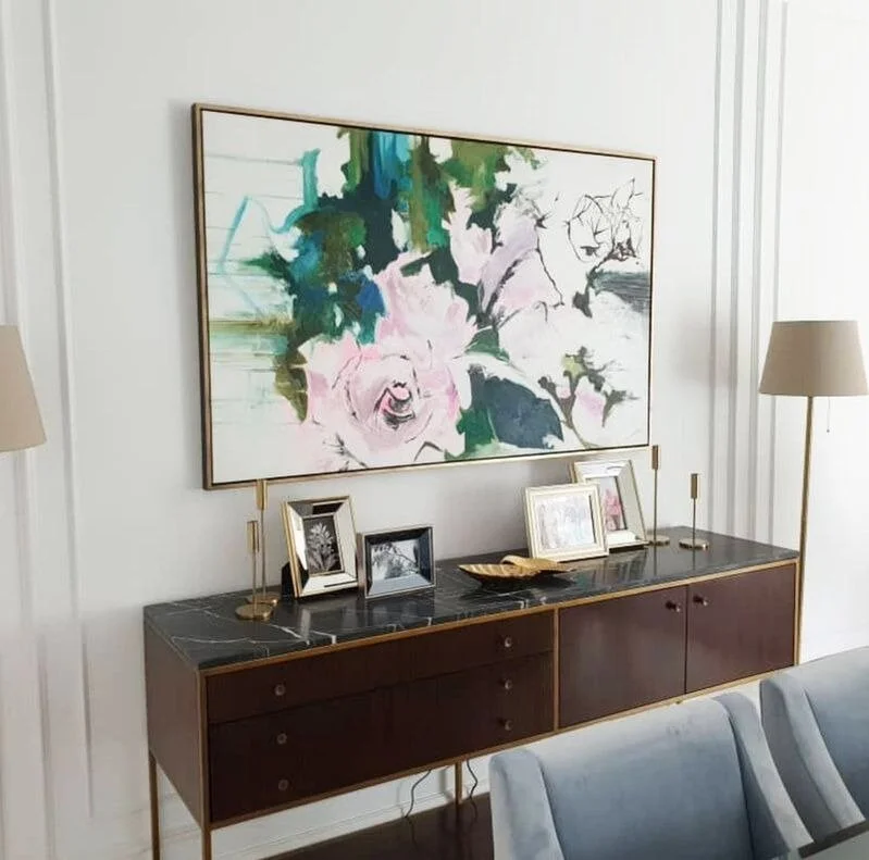 A mid-century modern sideboard with a black marble top, decorated with framed black-and-white photographs, a gold tray, and two gold candlesticks. Above it hangs an abstract watercolor painting of flowers in pink, green, and blue tones. Two cream-colored lamps with gold bases stand on either side of the sideboard in a white room.