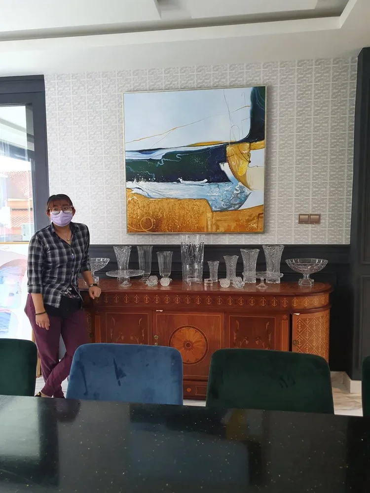 A woman wearing a face mask, glasses, plaid shirt, and purple pants stands next to a wooden sideboard with glassware on top, in a room with a large abstract painting on the wall.