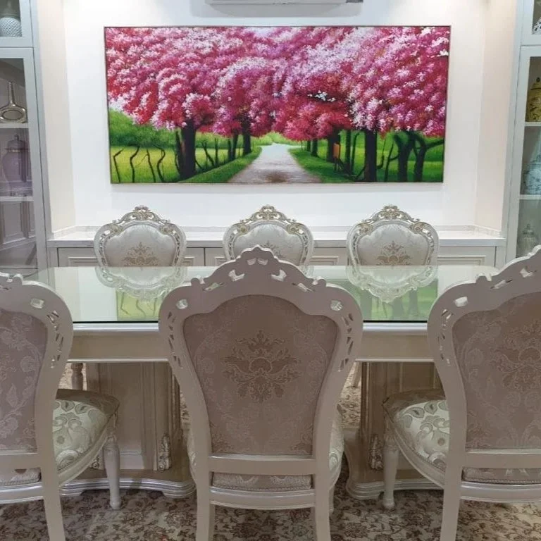 Dining room with a glass-top table, ornate white chairs with beige upholstery, a large painting of pink blossoms and a path on the wall, and decorative items on the shelves.