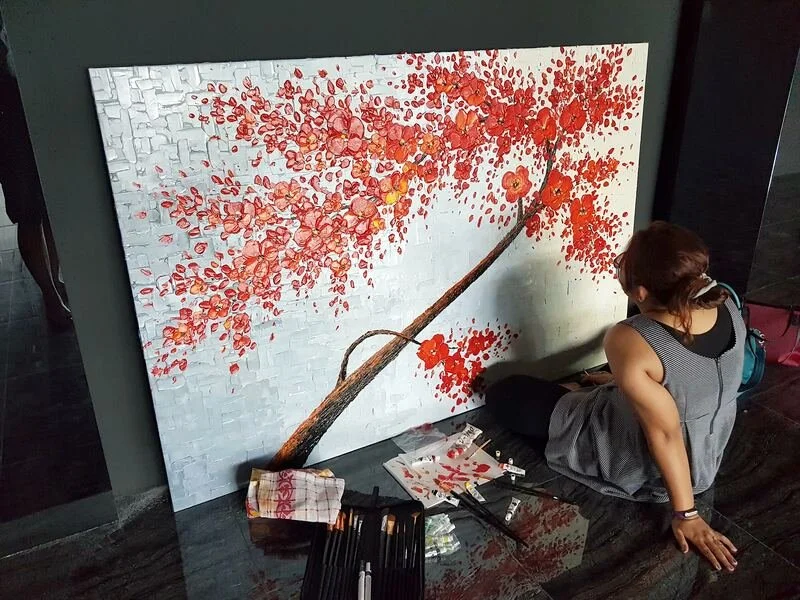 An artist working on a large painting of a cherry blossom tree with a white background, using various brushes and supplies on the floor.