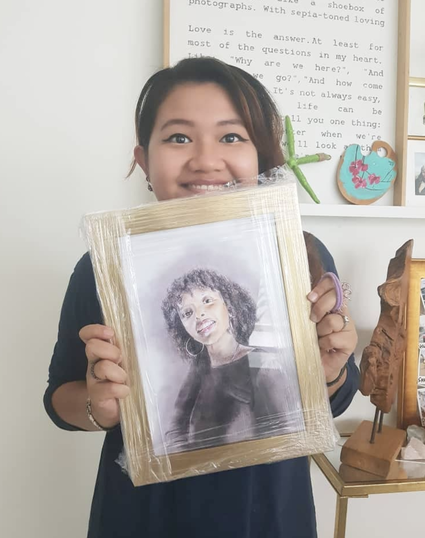A woman smiling and holding a framed pencil sketch of herself against a classroom wall decorated with a blue cloud-shaped note and a text poster.