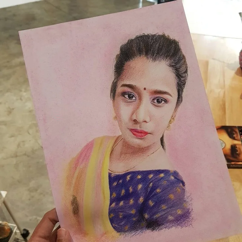 A crayon or pastel drawing of a young woman with dark hair tied back, wearing a traditional outfit with a yellow and pink dupatta over a navy blue dress with gold patterns, set against a pink background.