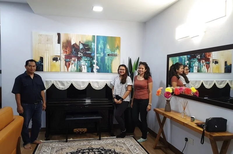 Four people standing in a living room next to a black piano, with paintings on the wall behind them, a large mirror on the right wall, a wooden console table with colorful flowers, and a decorative rug on the floor.
