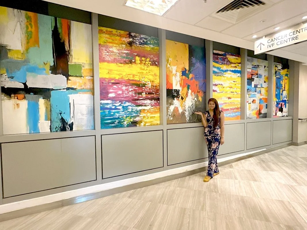 A woman in a floral outfit standing in front of a colorful abstract painting display in a hallway, with a sign pointing to the Cancer Centre.