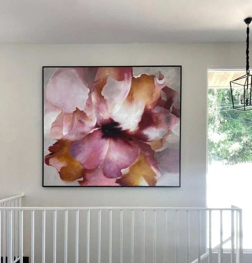 Large abstract floral painting with pink, purple, white, and brown tones hanging on a white wall next to a staircase railing and window showing greenery outside.