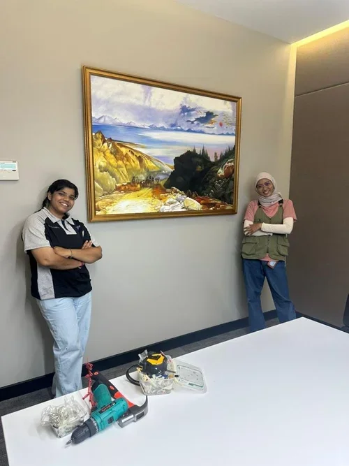 Two women standing in front of a framed landscape painting on a beige wall in a room with a white table. One woman is wearing a navy and white uniform with glasses, and the other is wearing a pink long sleeve shirt, a beige vest, a headscarf, and gla