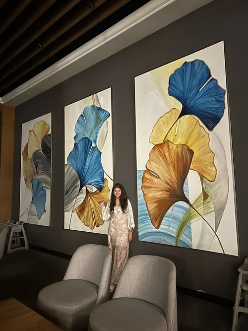 A woman standing in front of three large, colorful abstract paintings of ginkgo leaves on a dark gray wall in an art gallery or museum.