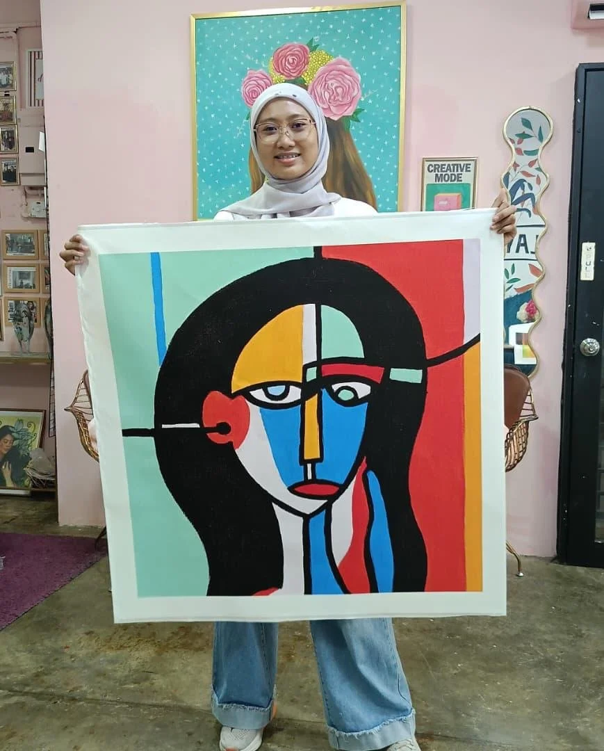 A woman wearing a white hijab and glasses is holding a colorful abstract portrait painting of a woman's face. The background has pink and teal decor, with a framed floral picture behind her.