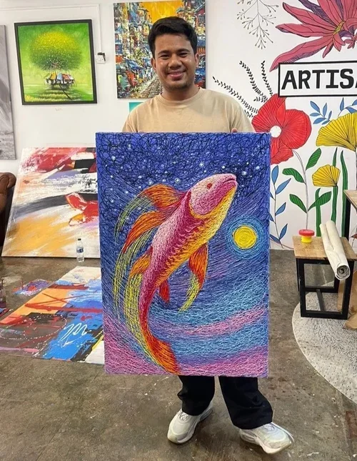A young man holding a colorful painting of a koi fish under a starry sky, with a yellow moon. The background has vibrant blue, pink, and purple hues, and there are various artworks and paintings displayed on the walls around him.