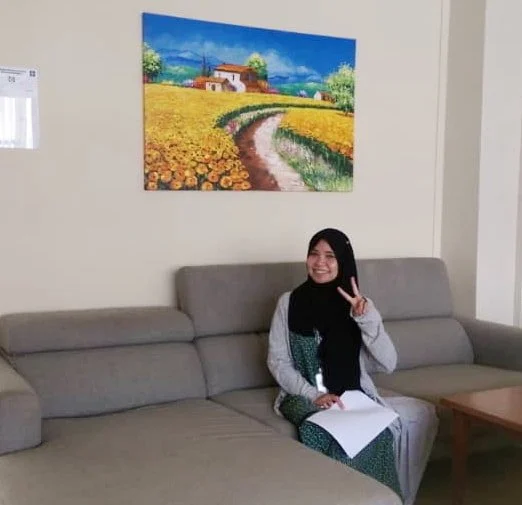 A woman wearing a hijab and gray dress sitting on a gray sofa, holding a paper and making a peace sign with her right hand, in a room with a colorful landscape painting on the wall.
