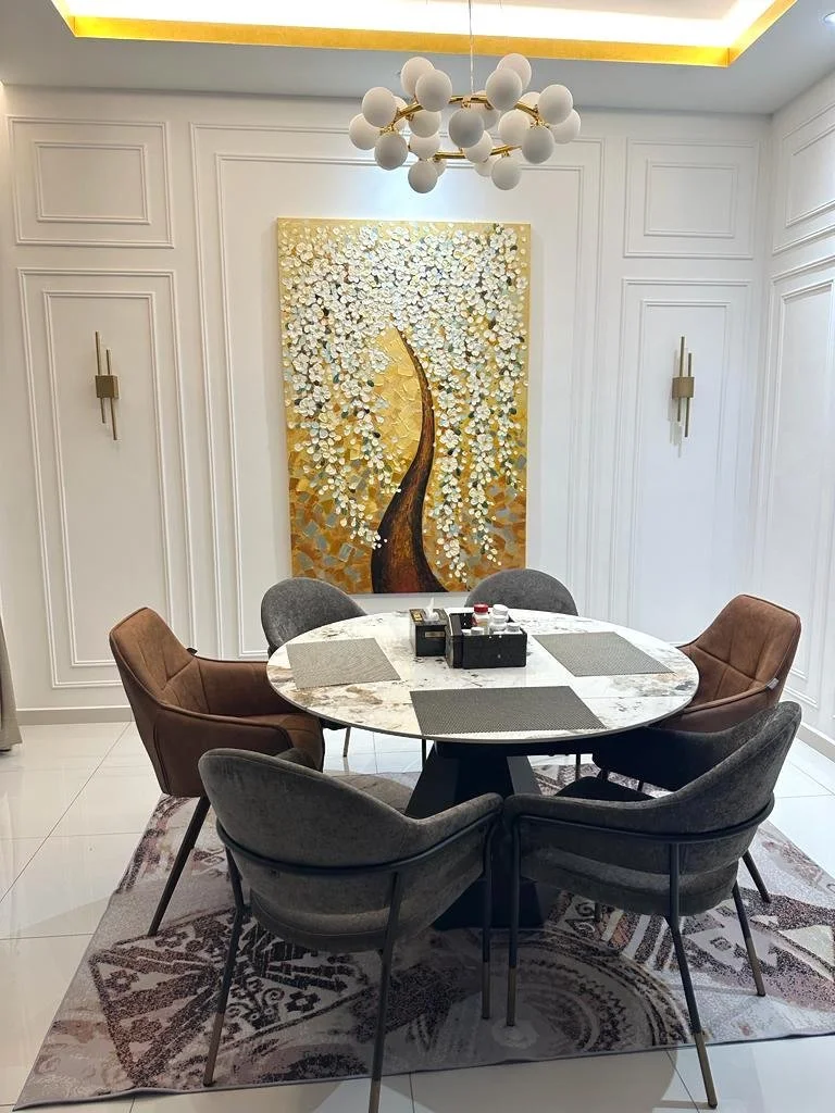 Modern dining room with a round marble table surrounded by six upholstered chairs in brown and gray. A large, textured gold and white floral painting hangs on the wall behind, with gold wall sconces on each side. A contemporary chandelier with white 