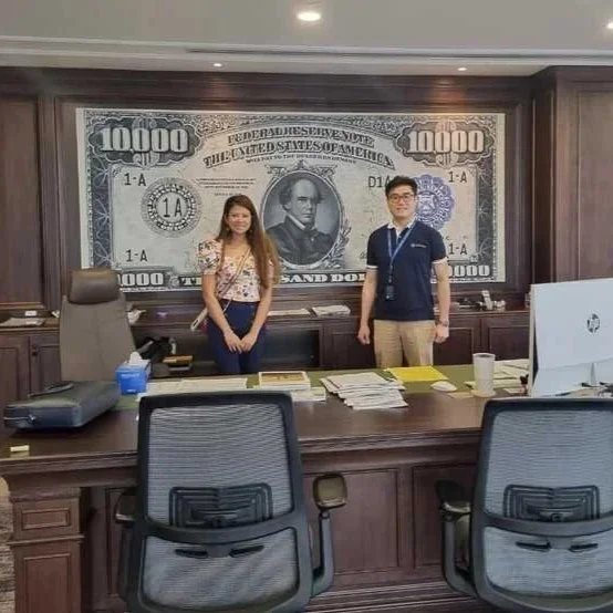 Two people standing in front of a large wall mural of a 10,000 dollar bill in an office setting.
