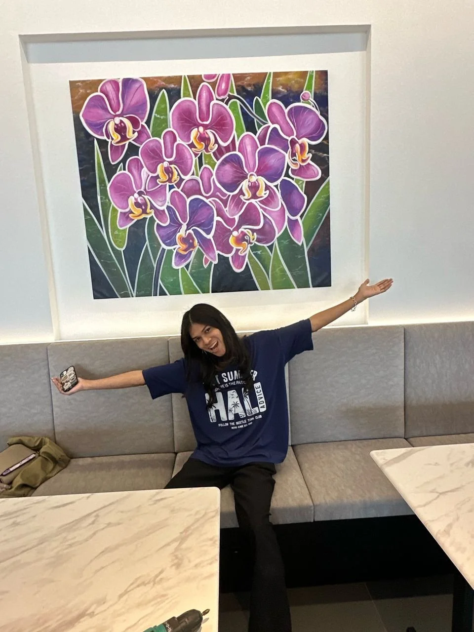 A woman sitting on a beige bench with her arms outstretched, smiling, in front of a large colorful painting of purple orchids on a white wall.