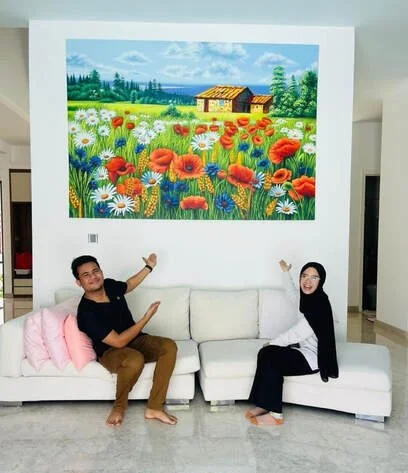Two people sitting on a white sofa in a living room, pointing at a large colorful painting of a field with flowers, trees, a house, and a blue sky hanging on the wall behind them.
